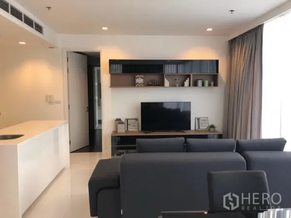 Condominium for rent in Sathon, Bangkok - Cozy living room with sectional sofa facing a TV console and wall shelves, under recessed lighting.