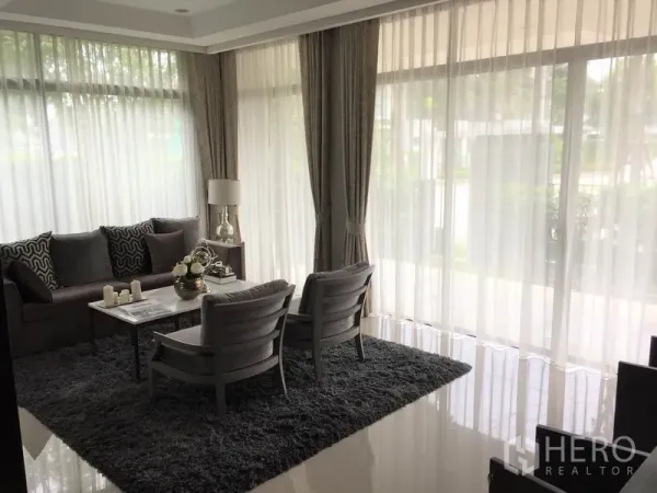 Detached House for rent or sale in Bang Kapi, Bangkok - Bright living room with floor-to-ceiling windows, sheer curtains and a grey sofa set.