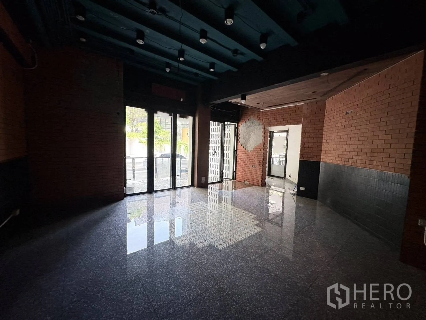 Retail Space for rent in Watthana, Bangkok - Street-facing retail interior with floor-to-ceiling glass doors, exposed brick walls, glossy stone flooring, and a dark ceiling with spotlights.