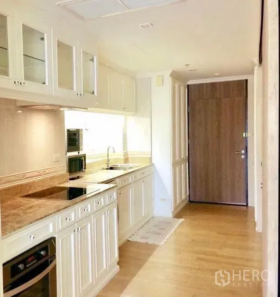Condominium for rent in Khlong Toei, Bangkok - Kitchen corridor with cooktop, oven, stone countertops, glass cabinets and warm wood flooring near the entry door.