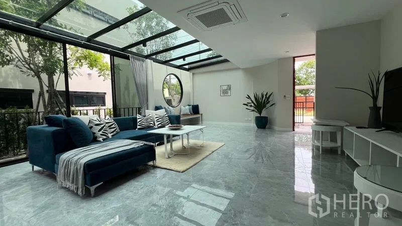 独立式住宅 出租在Lat Krabang, Bangkok - 客厅配蓝色沙发、圆窗与玻璃顶棚，景观花园。