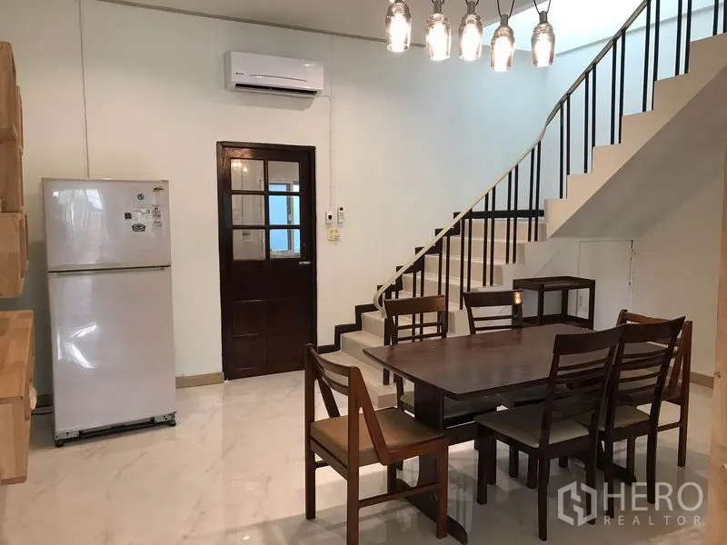 Townhouse for rent or sale in Phra Khanong, Bangkok - Dining area with wooden table, hanging lights, refrigerator and staircase with black railing.