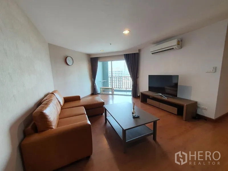 คอนโด ให้เช่าในHuai Khwang, Bangkok - ห้องนั่งเล่นโทนอบอุ่น โซฟาหนังรูปตัว L ทีวีจอแบน และประตูบานเลื่อนไปยังระเบียงชมวิวเมือง