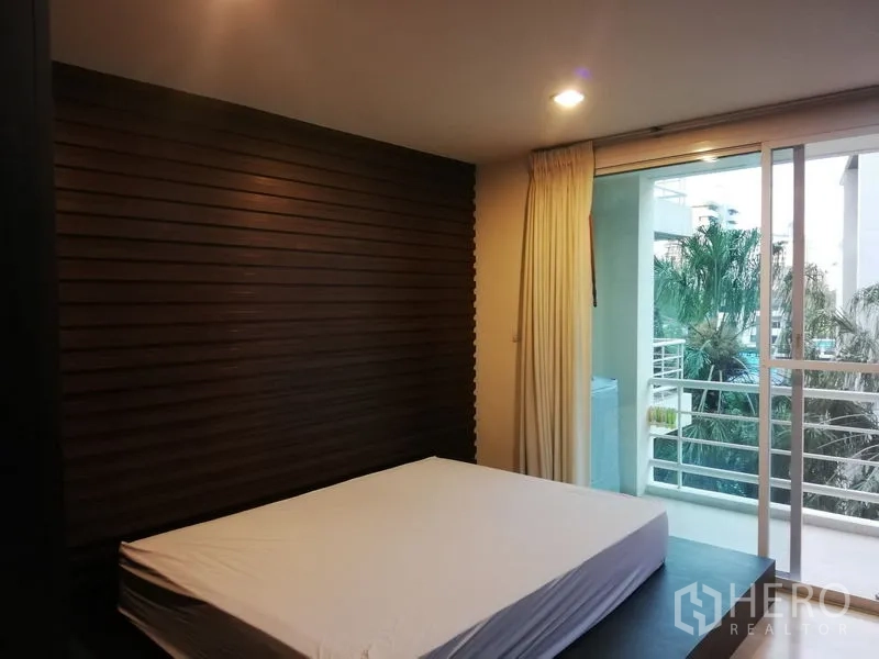 公寓 出租在Watthana, Bangkok - 主卧木质装饰墙，推拉门通阳台，可见树影与泳池区。