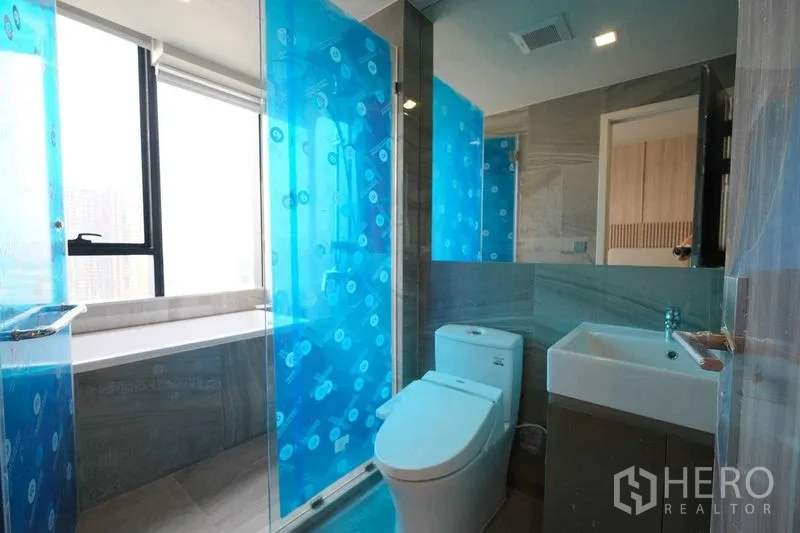 Condominium for rent in Khlong Toei, Bangkok - Contemporary bathroom with glass shower, bathtub by the window, toilet, and vanity.