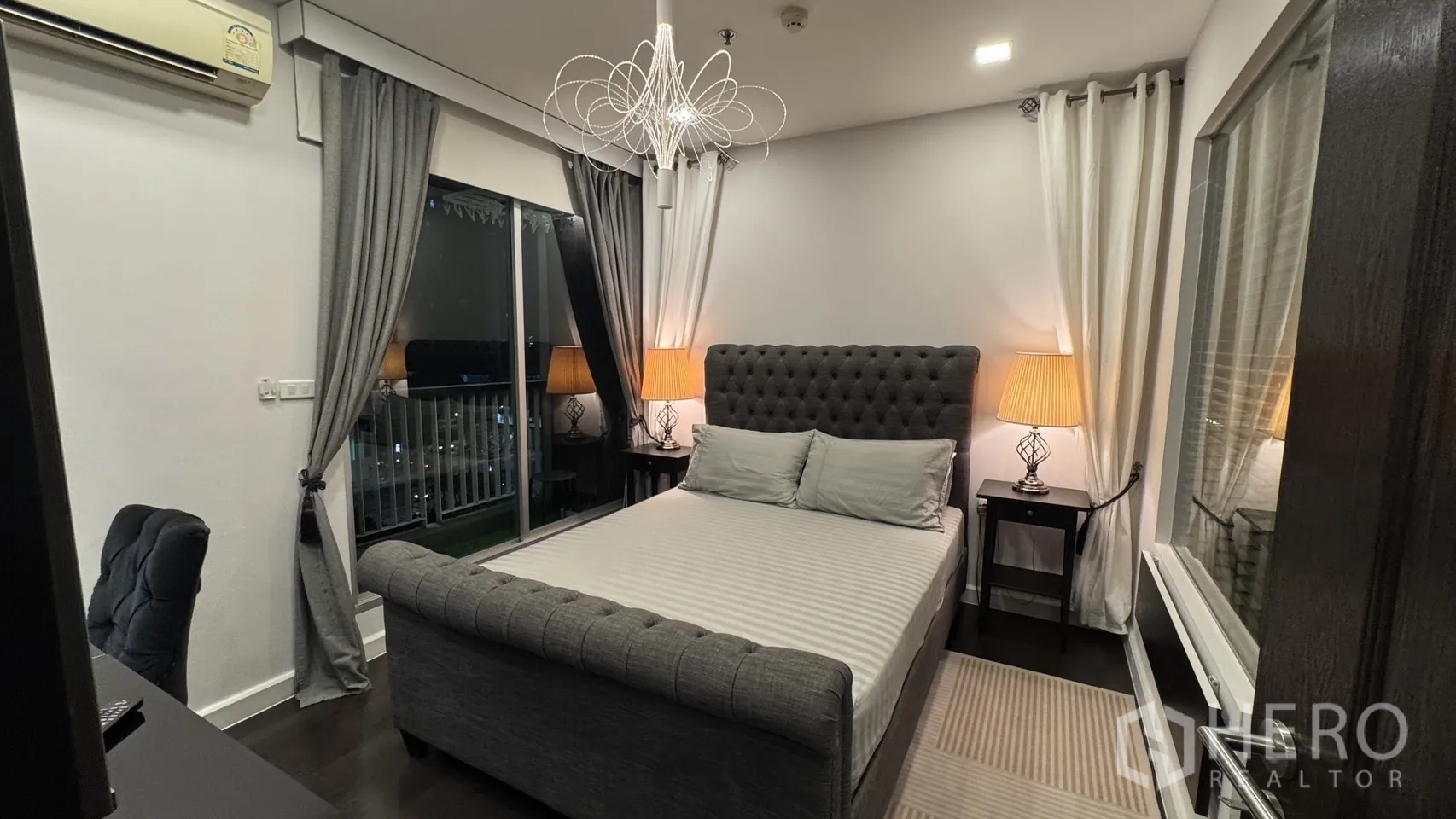Condominium for rent or sale in Khlong Toei, Bangkok - Bedroom with tufted grey bedframe, bedside lamps and access to a balcony.