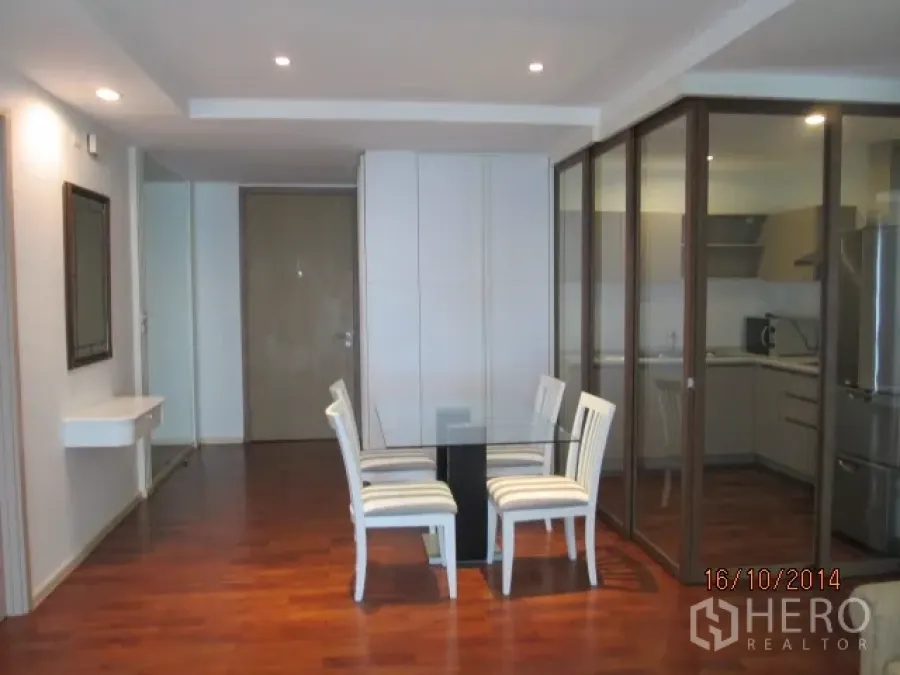 Condominium for rent or sale in Khlong Toei, Bangkok - Dining area with a glass table and white chairs beside a glass-partitioned kitchen, warm wood floors and recessed lighting.