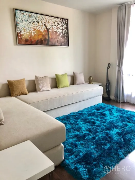 Detached House for rent in Bang Kapi, Bangkok - Cozy family room with long beige sofa, teal shag rug, wall art and a large curtained window.
