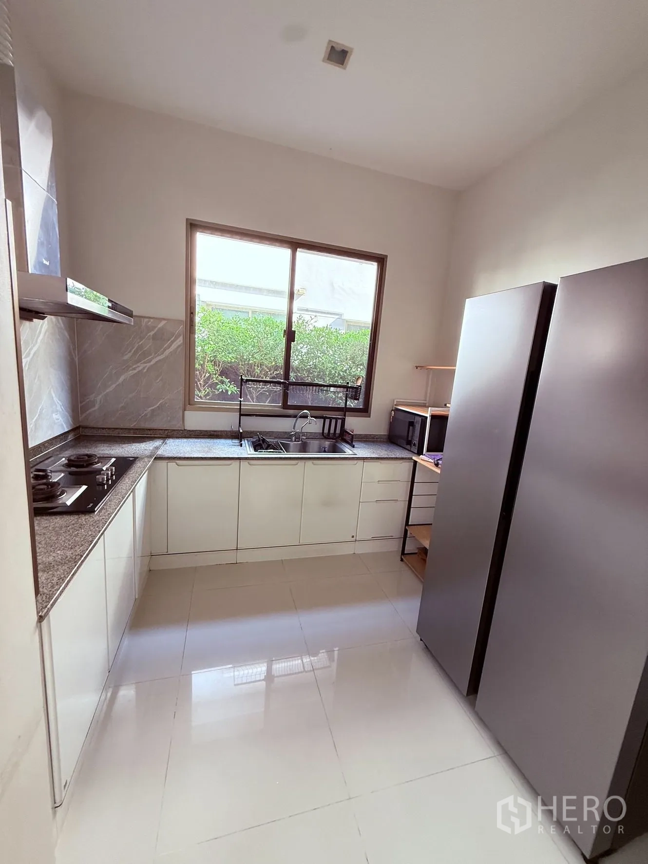 Detached House for rent or sale in Prawet, Bangkok - Bright kitchen with L-shaped countertop, gas hobs, sink under a window, and double-door fridge.