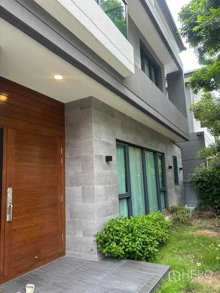 独立式住宅 出租在Mueang Nonthaburi, Nonthaburi - 入户区配实木门、灰色石材立面与绿篱花园。