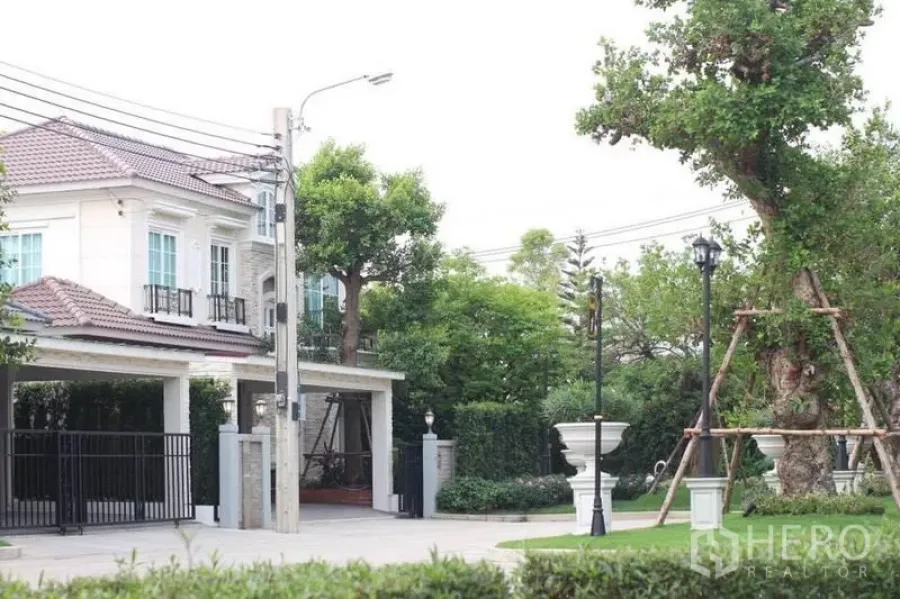 Detached House for rent or sale in Lat Phrao, Bangkok - Two-storey detached house with tiled roof, gated two-car carport and landscaped garden in a Bangkok neighborhood.