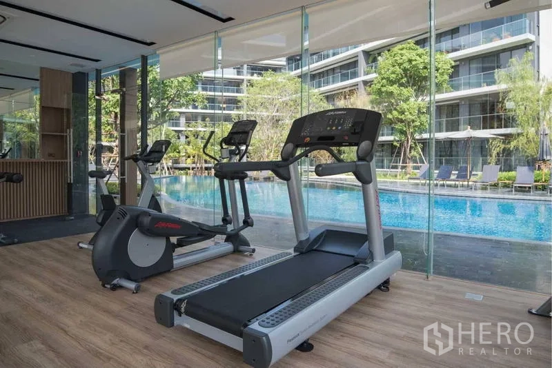 Condominium for sale - Glass-walled fitness room with treadmills overlooking a large swimming pool and greenery.