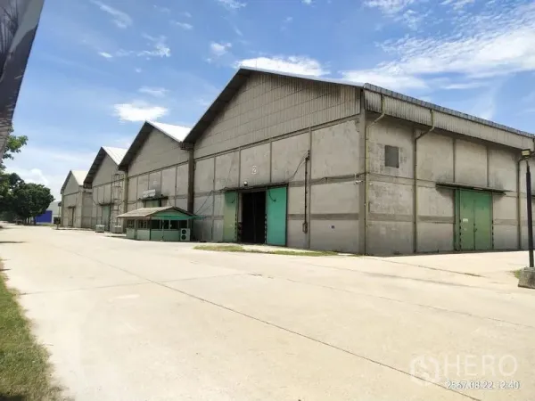 Warehouse for rent in Mueang Pathum Thani, Pathum Thani - Row of gable‑roof warehouses with green sliding doors and a wide concrete driveway under blue sky in Pathum Thani.