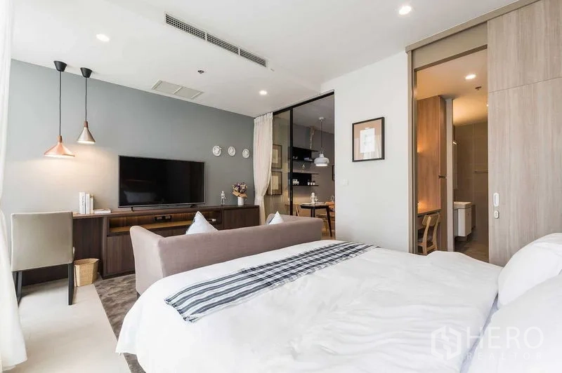 Condominium for rent in Pathum Wan, Bangkok - Bedroom view toward the TV wall and work desk with a glass door leading to the kitchen and pendant lights.