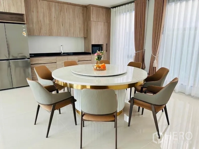 Detached House for rent or sale in Bang Kapi, Bangkok - Bright dining and kitchen area with six‑seat round marble table, wood cabinetry, and floor‑to‑ceiling curtains.