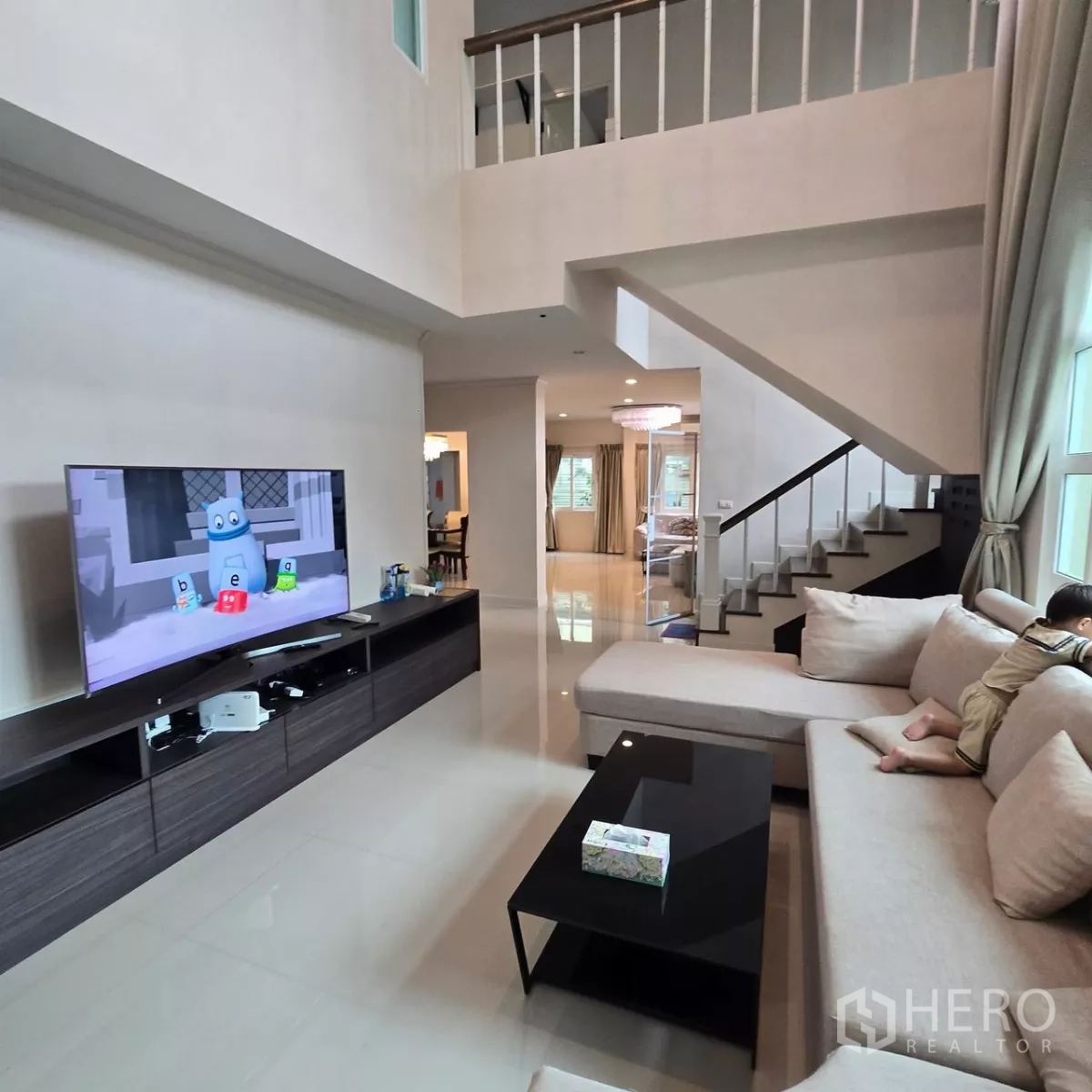 Detached House for rent in Mueang Pathum Thani, Pathum Thani - Living room with L-shaped sofa, TV wall and staircase view.