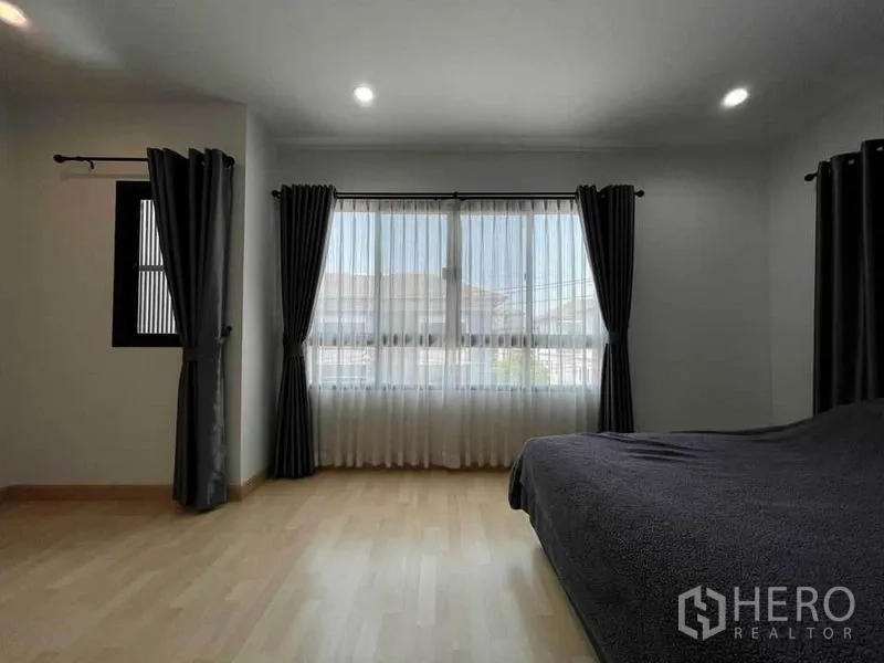 Townhouse for sale in Wang Thonglang, Bangkok - Spacious bedroom with large window, sheer curtains and dark drapes, light wood flooring and recessed lighting.