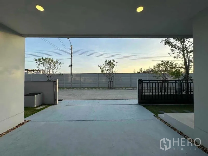 Detached House for rent in Bang Phli, Samut Prakan - Covered carport with gated driveway opening to a quiet street and landscaped verge at sunset.