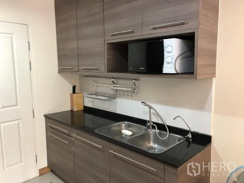 Condominium for rent in Khlong Toei, Bangkok - Modern kitchenette with wood cabinets, black countertop, double sink, and microwave.