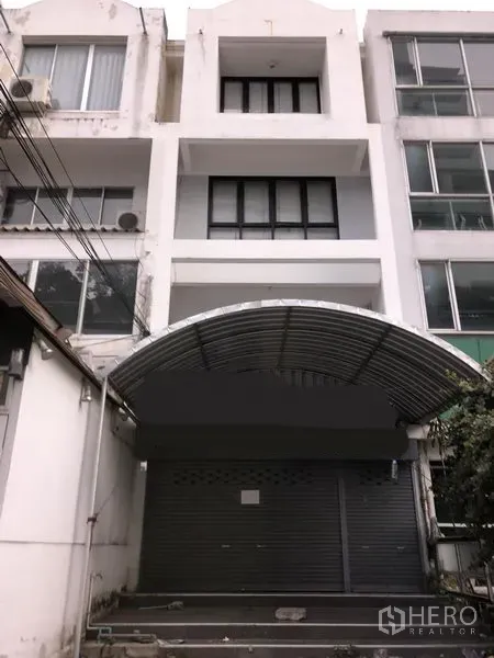 联排别墅 出售在Watthana, Bangkok - 曼谷瓦他那四层白色联排别墅外立面，带卷闸车库与弧形金属雨棚