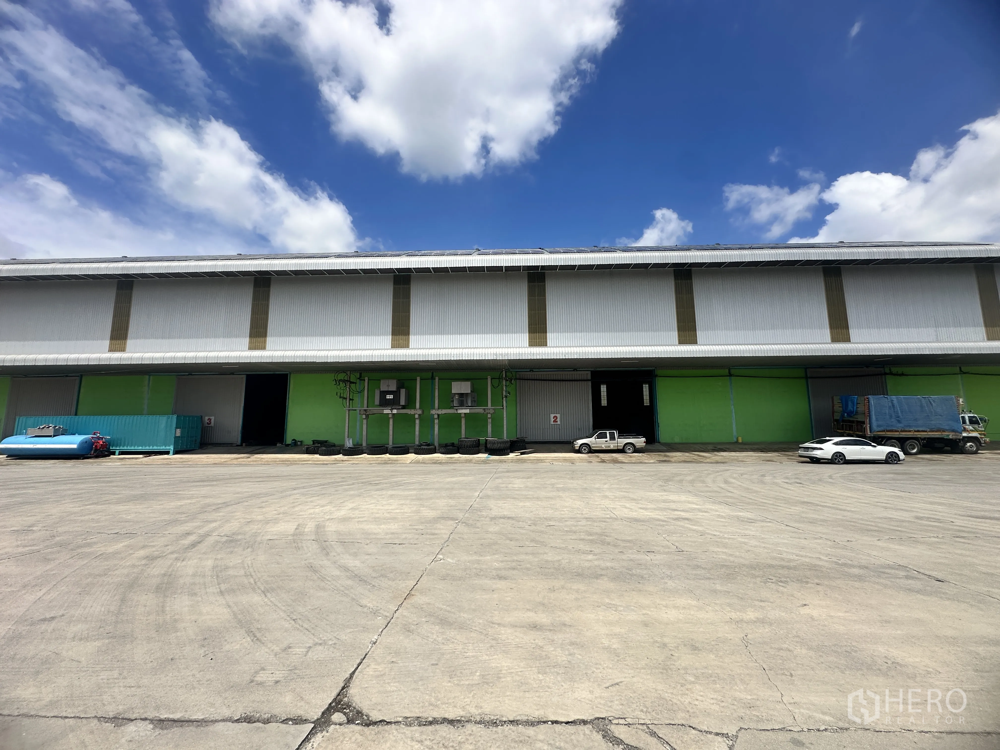 Warehouse for rent in Sai Noi, Nonthaburi - Front elevation of the warehouse featuring multiple loading bays and a wide paved yard with vehicles.