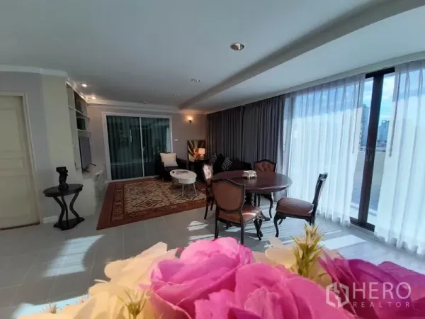 公寓 出租或出售在Watthana, Bangkok - 宽敞客厅，落地窗、餐桌、图案地毯与城市景观阳台，前景为鲜花。
