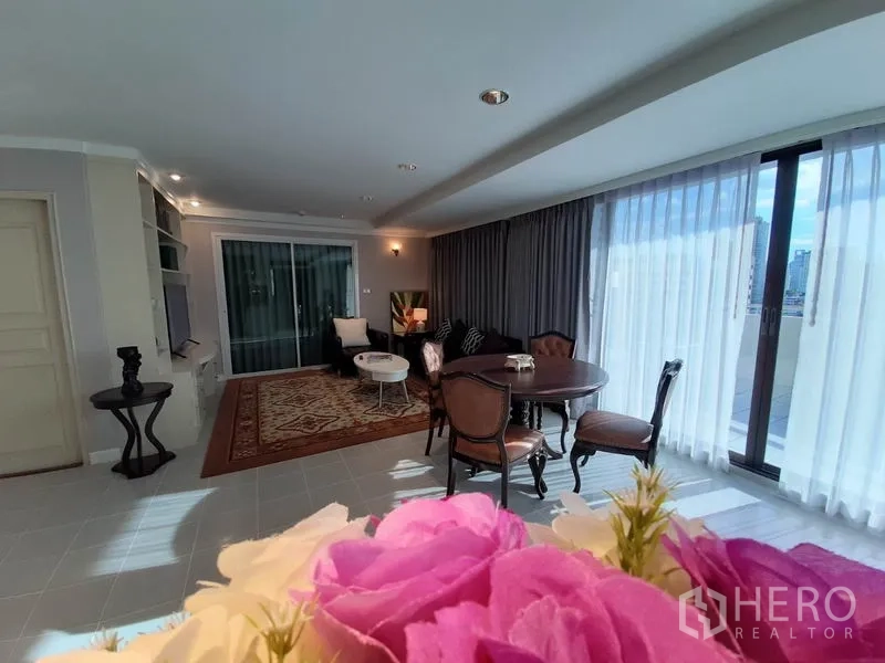 公寓 出租或出售在Watthana, Bangkok - 宽敞客厅，落地窗、餐桌、图案地毯与城市景观阳台，前景为鲜花。