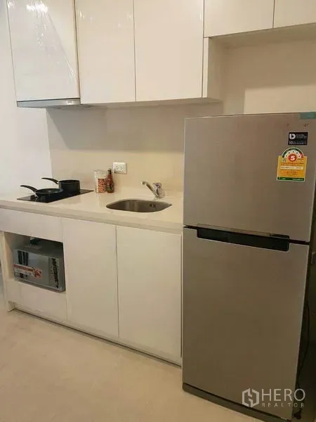 Condominium for rent in Khlong Toei, Bangkok - Compact kitchenette with glossy white cabinets, sink, electric hob and stainless refrigerator.