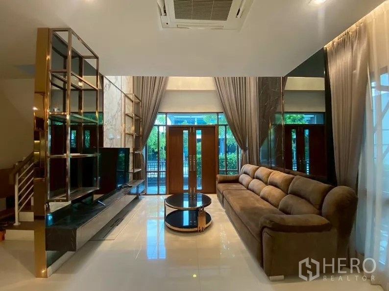 独立式住宅 出租在Prawet, Bangkok - 客厅采光充足，落地窗、大型沙发与金色置物架。