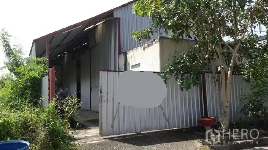 Warehouse for sale in Pathum Thani - Front view of warehouse with corrugated metal gate and tree shading the entrance.