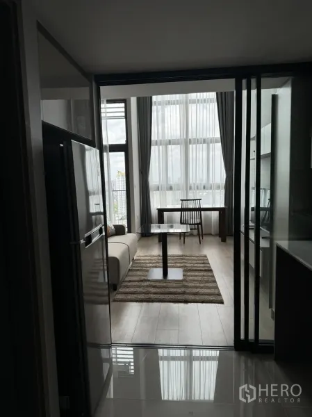 คอนโด ให้เช่าในHuai Khwang, Bangkok - ห้องนั่งเล่นคอนโดพร้อมหน้าต่างเต็มผนัง โซฟา พรม และโต๊ะทานอาหาร มองจากโซนครัว