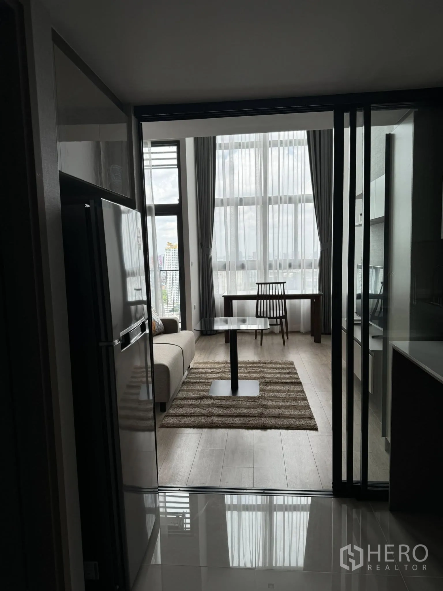 คอนโด ให้เช่าในHuai Khwang, Bangkok - ห้องนั่งเล่นคอนโดพร้อมหน้าต่างเต็มผนัง โซฟา พรม และโต๊ะทานอาหาร มองจากโซนครัว