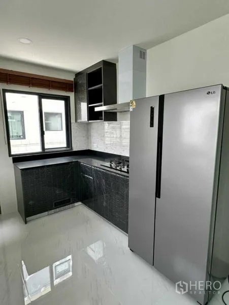 Detached House for rent in Bang Phli, Samut Prakan - Fitted L-shaped kitchen with black cabinets, gas hob, hood, and tall stainless refrigerator.