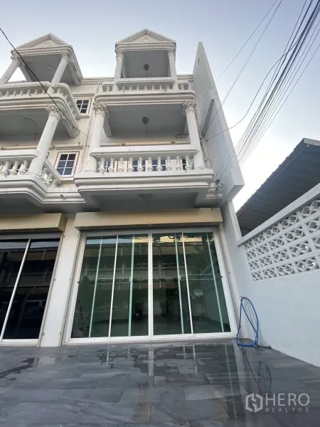 Townhouse for rent in Huai Khwang, Bangkok - Street view of a white three-storey townhouse with wide sliding glass doors and a spacious tiled forecourt.