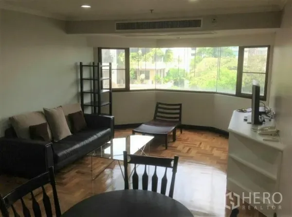 Condominium for rent in Watthana, Bangkok - Bright living room with curved bay windows, parquet wood floors, black leather sofa, glass coffee table, TV console, and leafy neighborhood view.