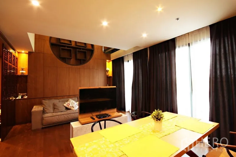 Condominium for rent or sale in Khlong Toei, Bangkok - Cozy living area with sofa, TV stand, floor-to-ceiling curtains, and a feature wood wall.