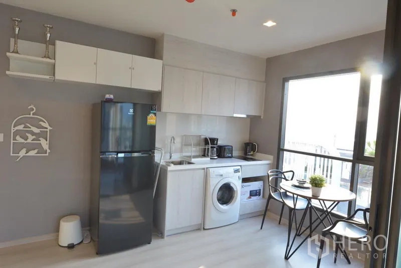 Condominium for sale in Khlong Toei, Bangkok - Modern kitchenette with full cabinetry, refrigerator, washing machine, and a two-seat dining table by the window.