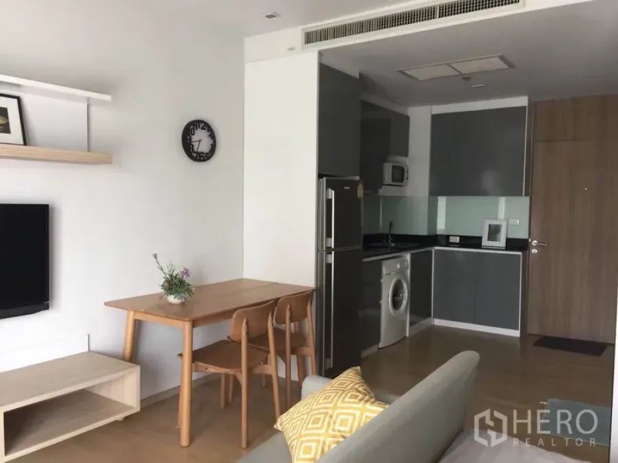 Condominium for rent or sale in Bangkok - Open-plan living area with wooden dining set and L-shaped kitchen featuring fridge, microwave and in-unit washer in a modern Sukhumvit condo near BTS Phrom Phong.