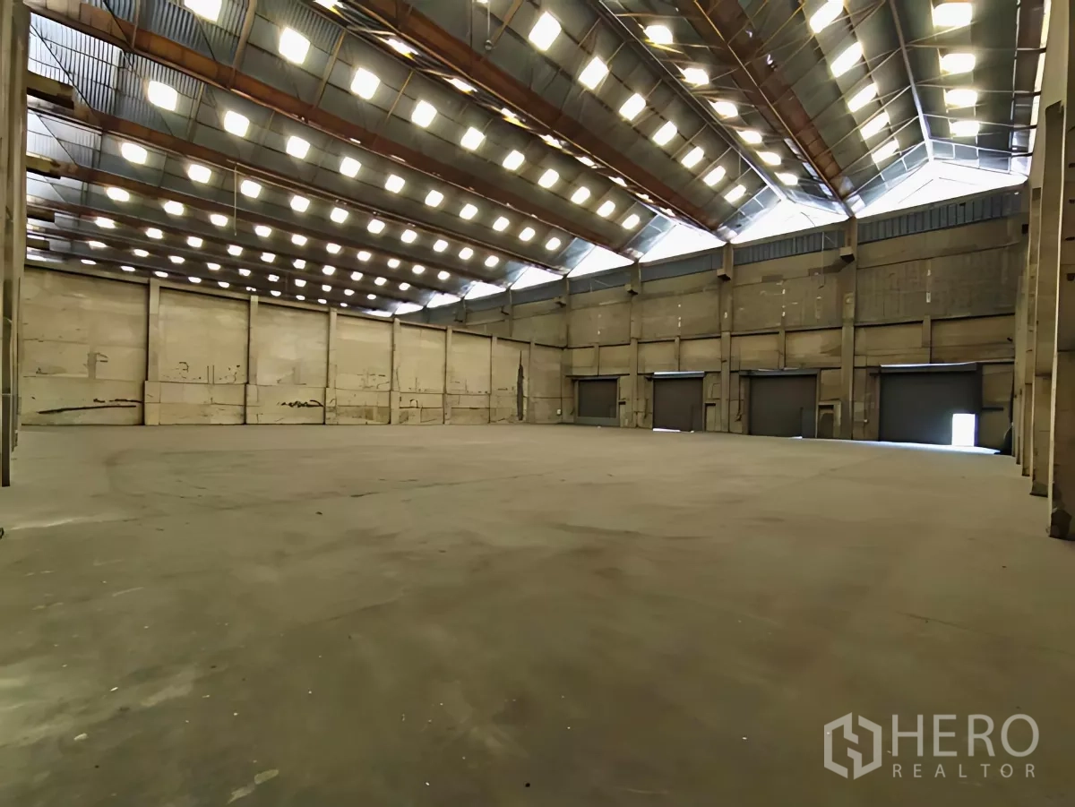 Factory for rent in Khlong Toei, Bangkok - Spacious factory interior with a high roof, skylights, concrete floor, and multiple roller shutter doors.