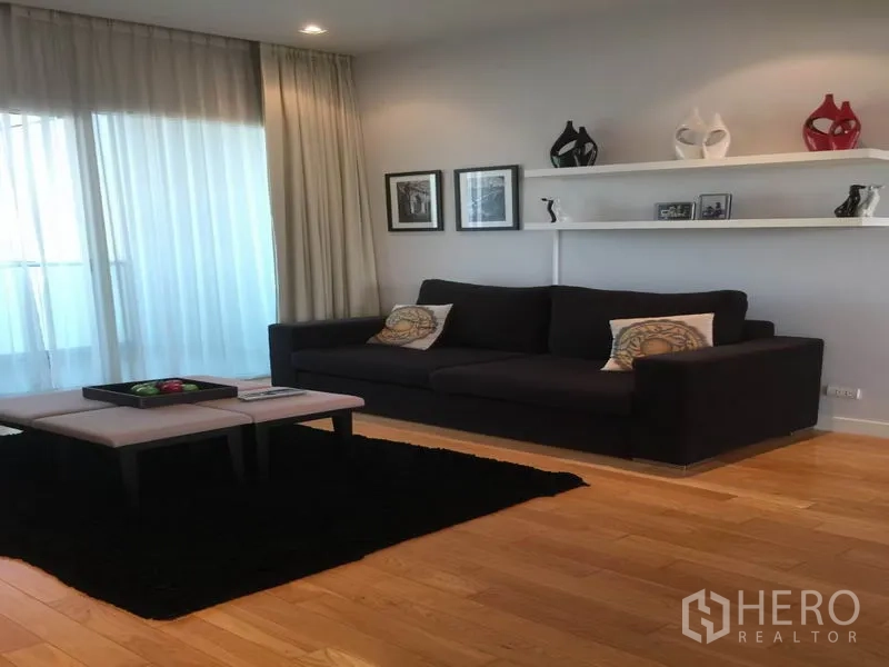 Condominium for rent in Khlong Toei, Bangkok - Living area featuring a dark sofa, wall shelves and coffee table beside floor‑to‑ceiling windows.