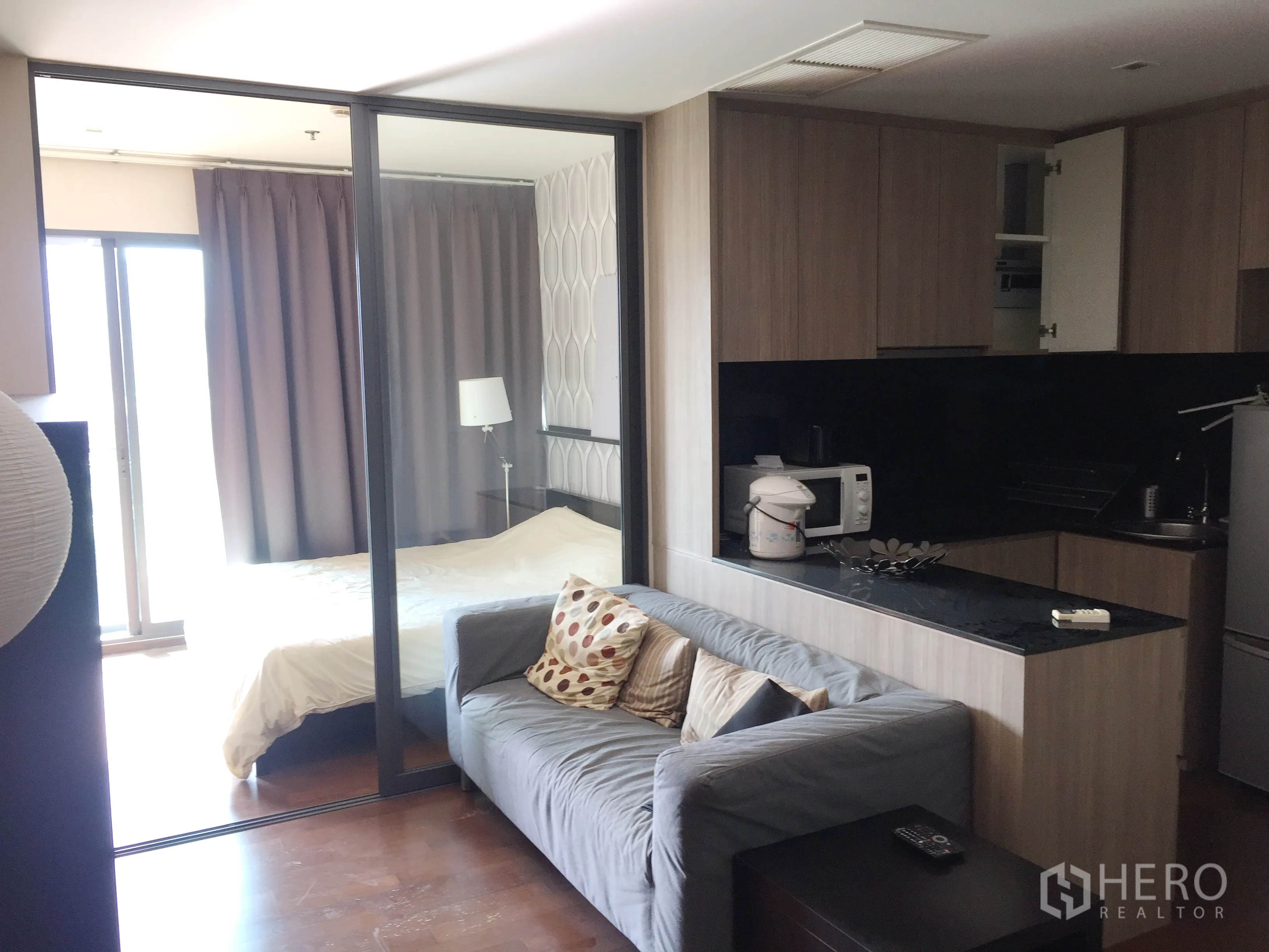 Condominium for rent in Khlong Toei, Bangkok - Open-plan living area with grey sofa, compact kitchen with black countertop, and a bedroom behind sliding glass.