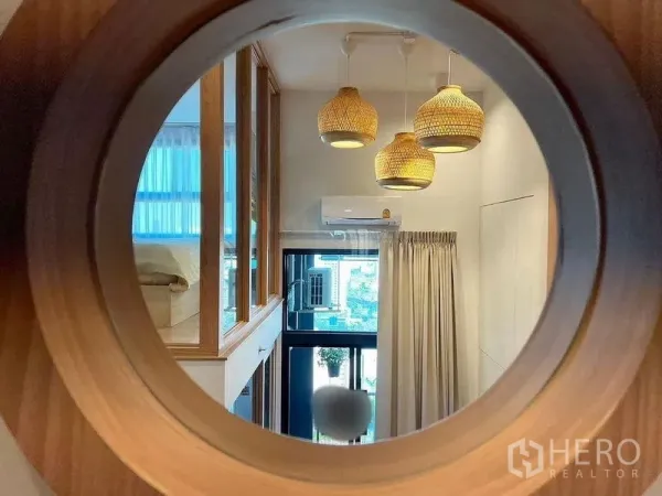 Condominium for rent in Huai Khwang, Bangkok - Circular wooden window framing a view of the living space with rattan pendant lights, curtains and air conditioner.