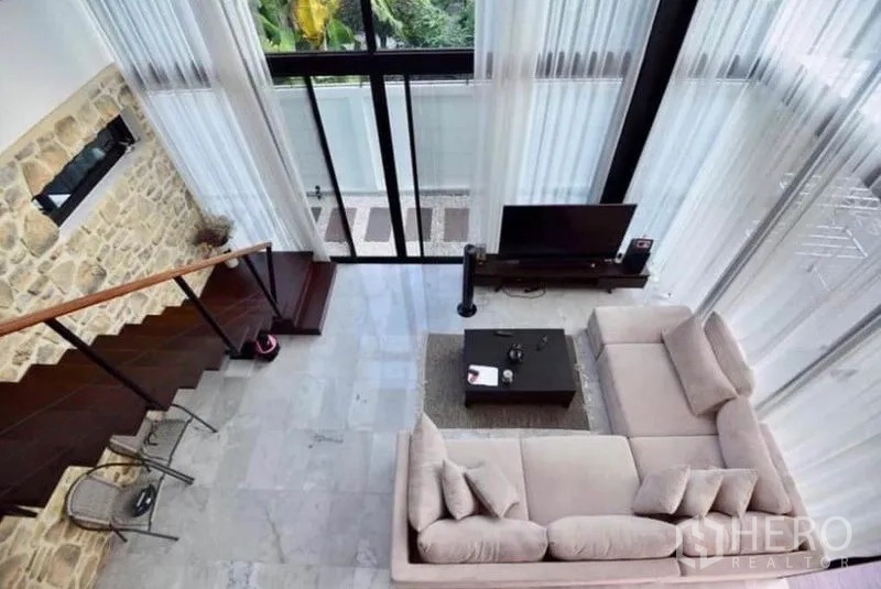 Townhouse for rent in Din Daeng, Bangkok - Double-height living room with floor-to-ceiling windows, L-shaped sofa, stone feature wall and staircase.