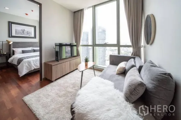 公寓 出租在Ratchathewi, Bangkok - 客厅配灰色沙发与电视柜，大窗采光充足，旁边通往卧室并享城市景色。