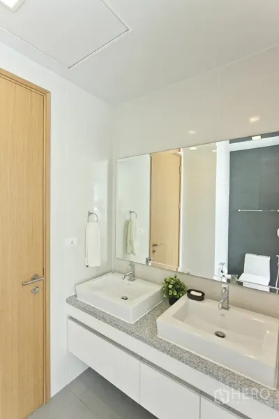 Condominium for rent in Khlong Toei, Bangkok - Second bathroom featuring double vanity sinks, large mirror, and light wood door.