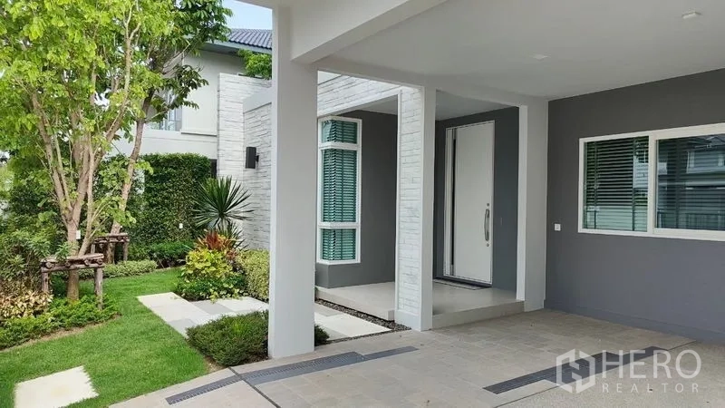 Detached House for rent in Prawet, Bangkok - Shaded front porch and carport leading to a large white entrance door beside trimmed greenery.