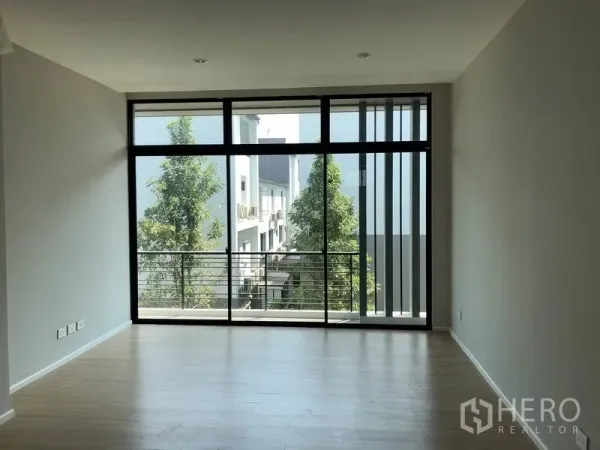 Townhouse for rent or sale in Saphan Sung, Bangkok - Spacious living area with floor-to-ceiling windows and a balcony overlooking trees.