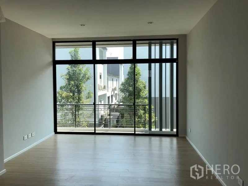 Townhouse for rent or sale in Saphan Sung, Bangkok - Spacious living area with floor-to-ceiling windows and a balcony overlooking trees.