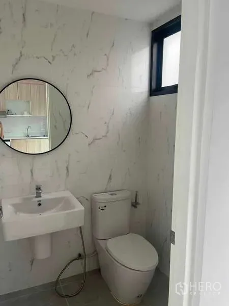 Townhouse for rent in Suan Luang, Bangkok - Compact bathroom with marble-effect walls, round mirror, square basin, toilet and a small window.
