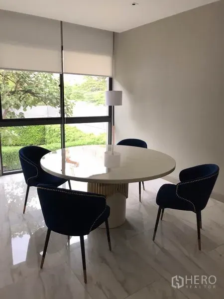 独立式住宅 出租在Watthana, Bangkok - 采光良好的餐区，白色圆桌与四把海军蓝椅，临大窗与百叶，地面为大理石。